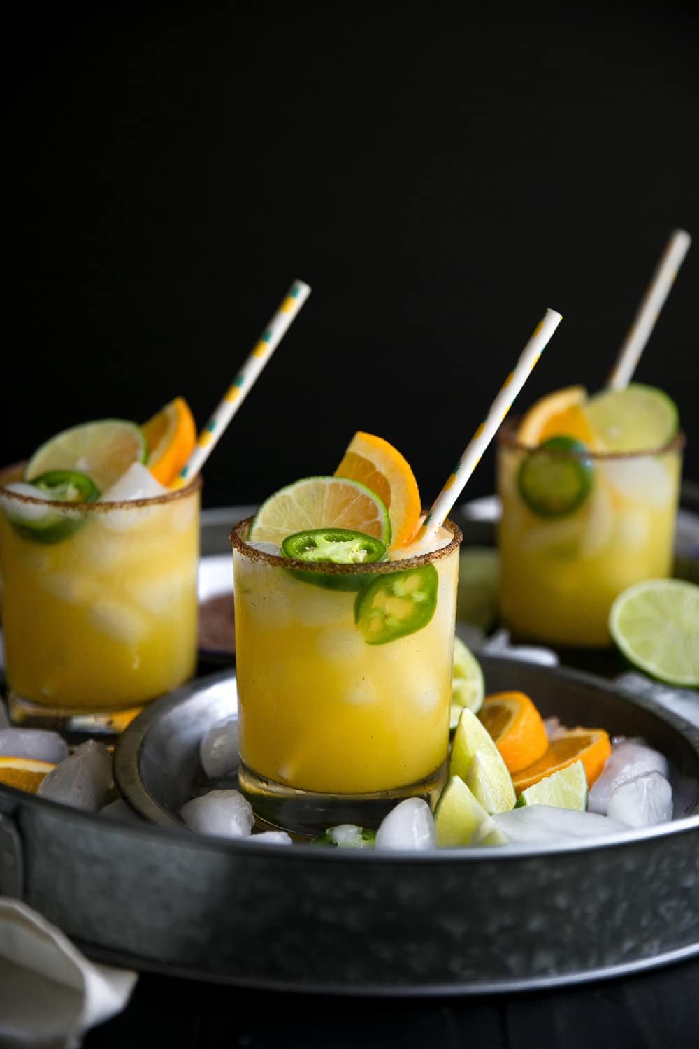 Three glasses filled with Spicy Orange Margarita and garnished with fresh jalapeno, lime, and orange slices with a sweet and spicy chili salt rim.