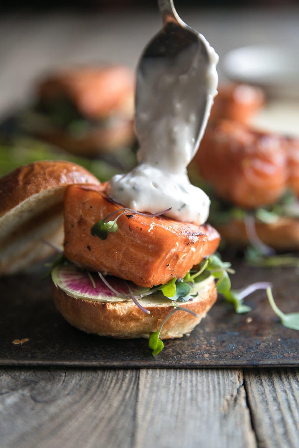 Grilled salmon fillet on the bottom half of a bun being topped with garlic aioli.