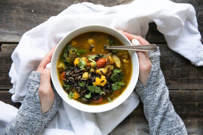 Hands holding a white bowl filled with nourishing turmeric broth soup filled with cailiflower, lentils, chickpeas, spinach, tomatoes, and wild rice.
