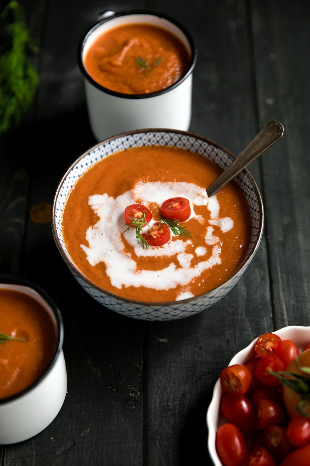 Soup bowl and two white mugs filled with roasted red pepper and tomato soup garnished with heavy cream and fresh cherry tomatoes.