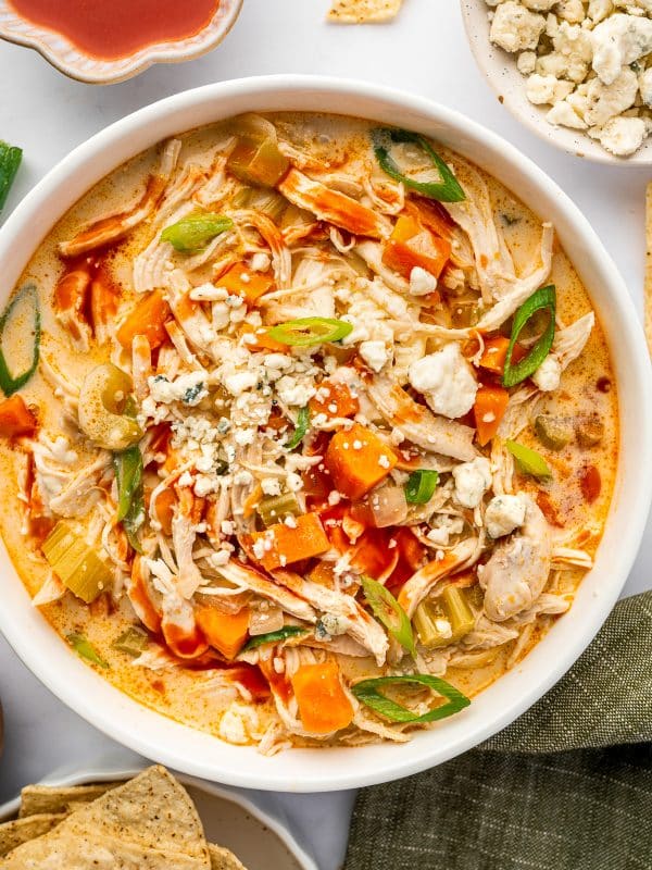 close-up of creamy buffalo chicken soup in a white bowl showing chunks of chicken, carrots, and celery and garnished with blue cheese, hot sauce, and green onions.