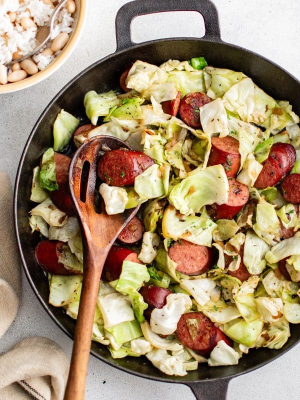 A skillet filled with kielbasa and cabbage, with a wooden spoon resting inside, served alongside a bowl of rice and beans.
