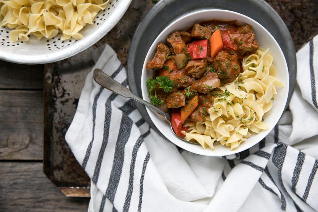 Bowl filled with beef goulash and egg noodles and set on a wood table.