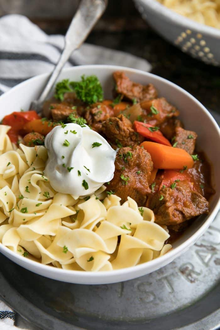Large white bowl filled with Hungarian goulash with thick chunks of beef, egg noodles, and topped with sour cream.