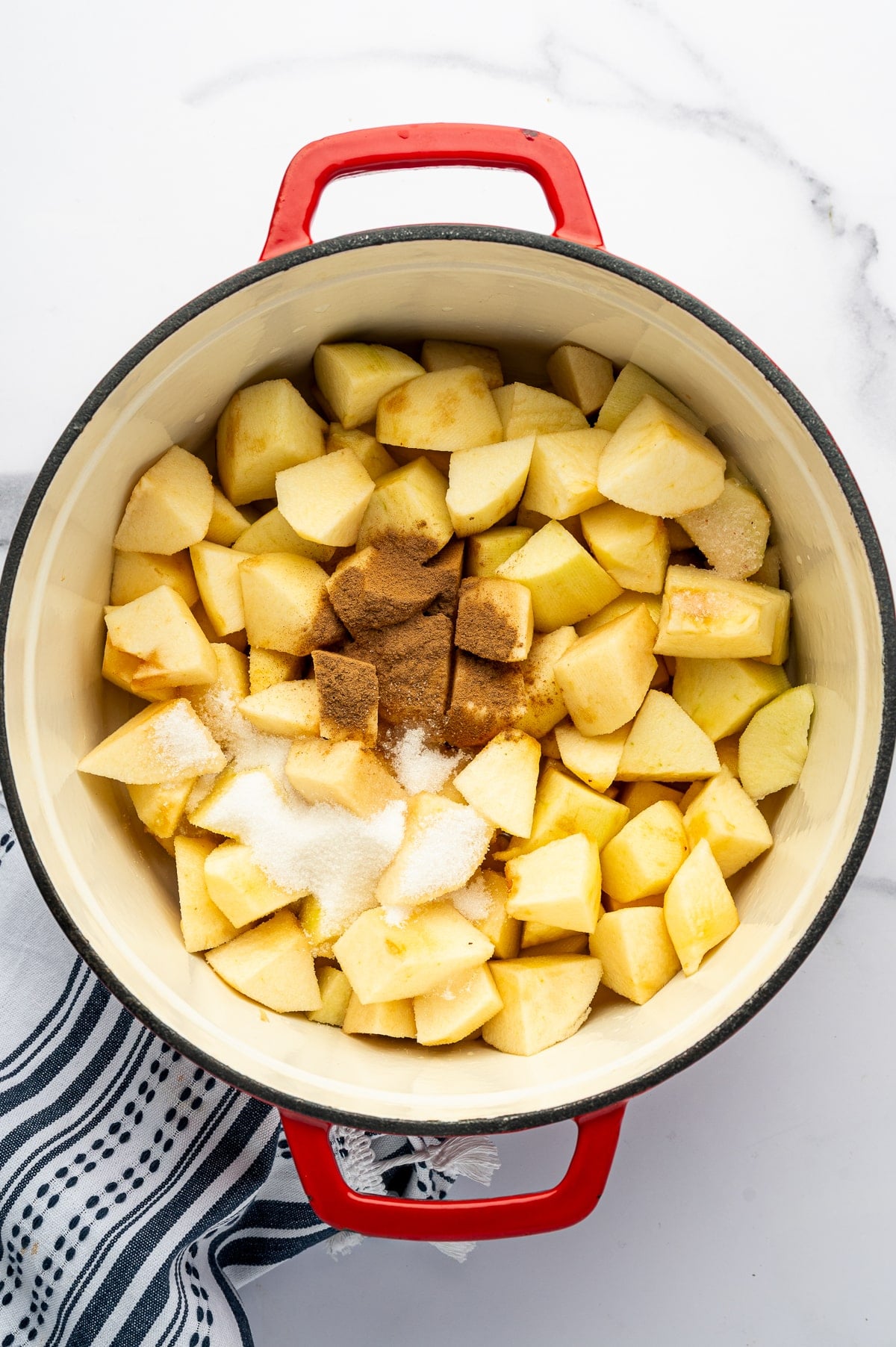Overhead view of peeled and chopped yellow apple pieces in a cream-colored enameled pot with bright red handles, topped with granulated sugar, ground cinnamon, and salt. A black-and-white striped kitchen towel is partially visible beside the pot on a white marble surface.