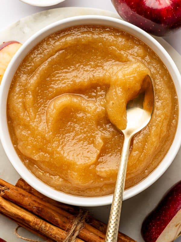 Overhead view of a round white ceramic bowl filled with smooth golden-brown applesauce, with a hammered gold-colored spoon lifting applesauce from the right side. A whole red apple, red apple wedges, a white bowl of ground cinnamon, and cinnamon sticks tied with twine surround the bowl on a pale stone surface.