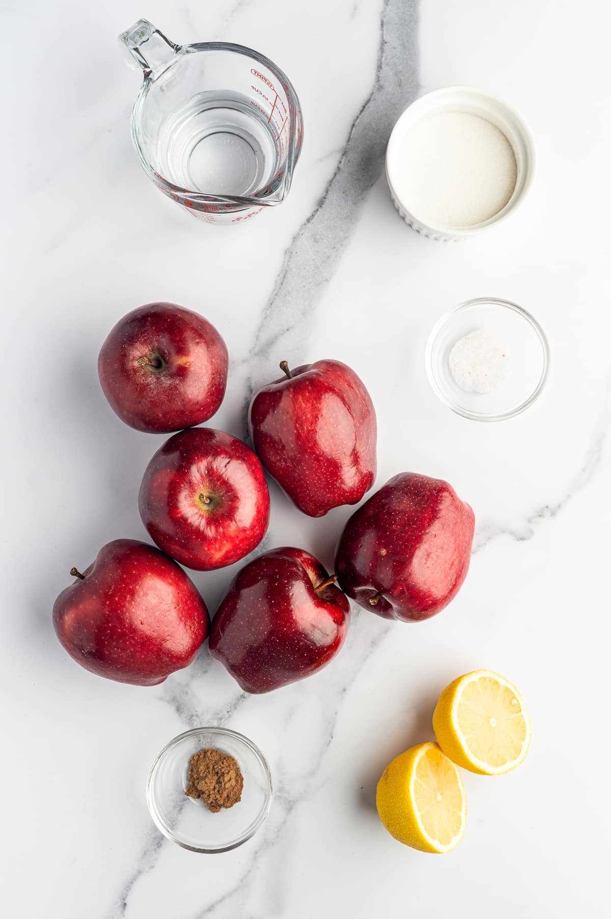 Overhead view of homemade applesauce ingredients on a white marble surface, including six red apples, a glass measuring cup filled with water, a white ramekin of granulated sugar, a small clear bowl of salt, a small clear bowl of ground cinnamon, and two lemon halves.