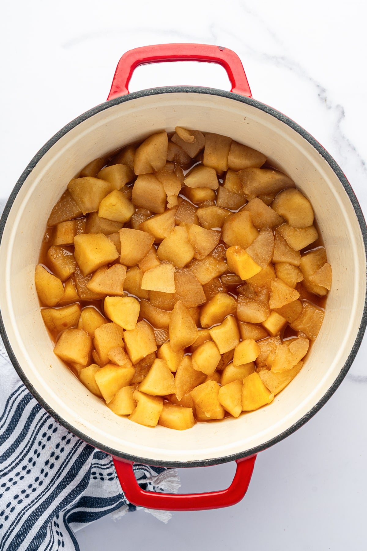 Overhead view of softened golden-brown apple chunks surrounded by cinnamon-spiced cooking liquid in a cream-colored enameled pot with bright red handles. A black-and-white striped towel sits beside the pot on a white marble surface.