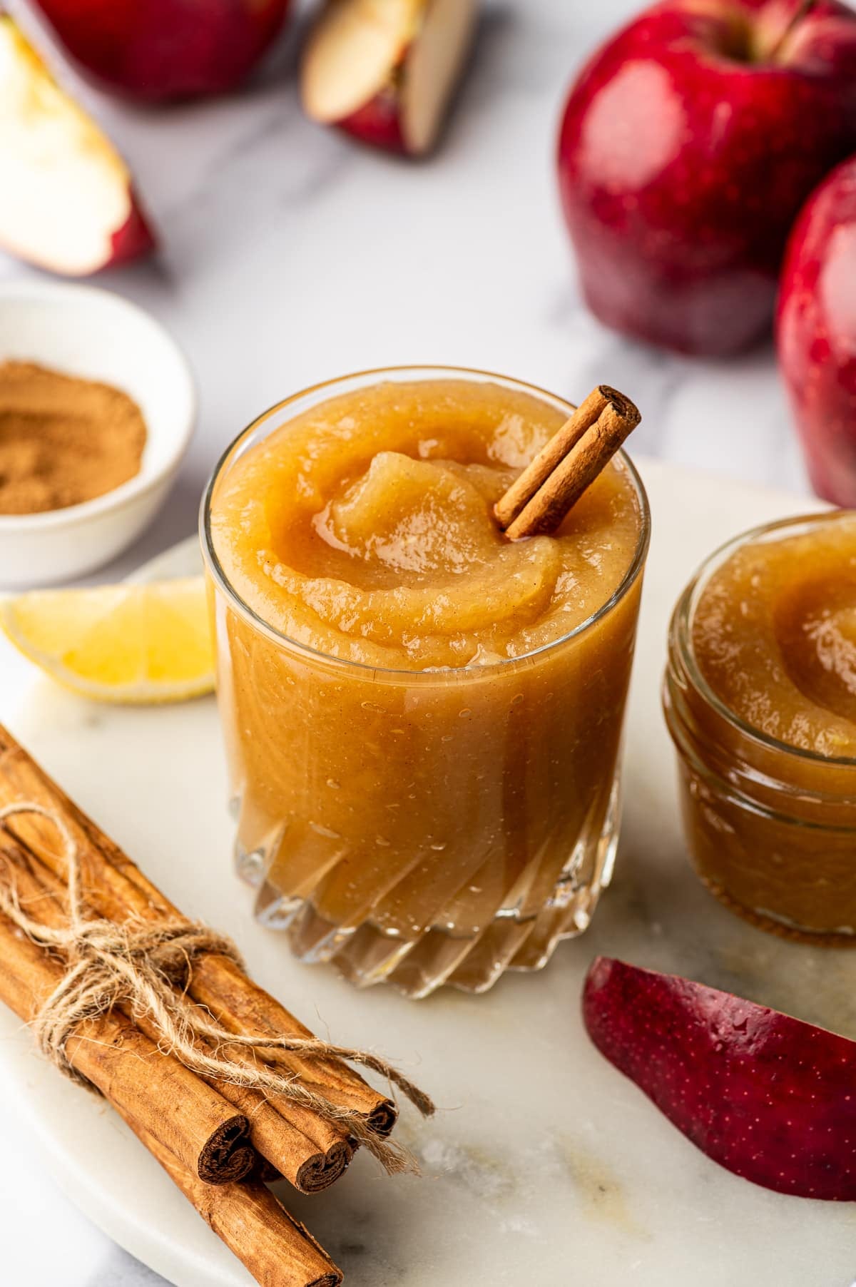 Clear faceted glass filled with thick golden-brown homemade applesauce and garnished with a cinnamon stick, with a small glass jar of applesauce to the right. Red apples, apple wedges, a lemon wedge, a white bowl of cinnamon, and cinnamon sticks tied with twine surround the glass on a pale stone board.