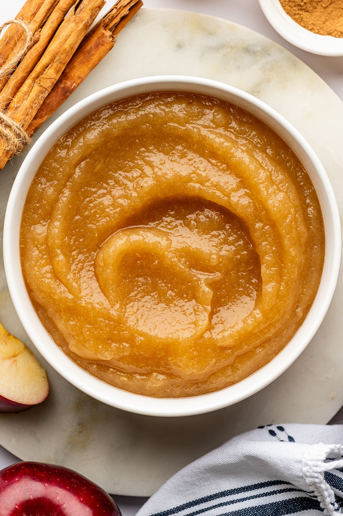 Overhead view of smooth golden-brown homemade applesauce swirled inside a round white ceramic bowl on a pale stone board. Cinnamon sticks tied with twine sit at the upper left, a small white bowl of ground cinnamon is at the upper right, a red apple and apple wedge appear along the bottom edge, and a white towel with dark blue stripes rests at the lower right.