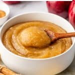 Pinterest collage for a homemade applesauce recipe featuring three photos across the top: thick golden-brown applesauce in a clear faceted glass garnished with a cinnamon stick, an overhead display of red apples with water, sugar, salt, cinnamon, and lemon, and applesauce in a white ceramic bowl with a wooden spoon lifting a scoop. Large black text on a white background reads "Homemade Applesauce Recipe" with a yellow line between the title lines. The bottom photo shows an overhead close-up of golden applesauce in a clear glass garnished with a cinnamon stick, with cinnamon sticks and lemon partially visible. "THEFORKEDSPOON.COM" appears in black text along the bottom.