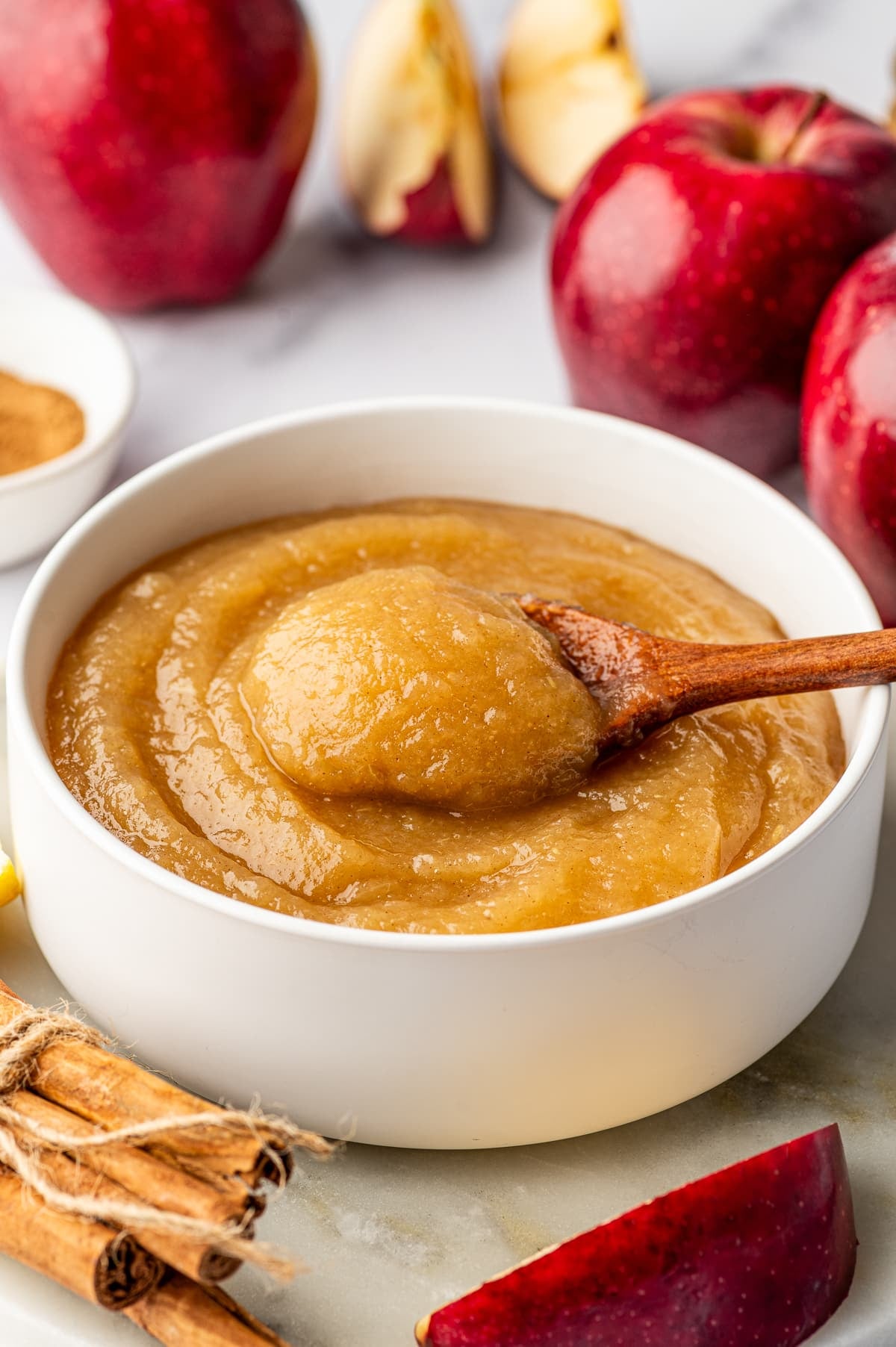 White ceramic bowl filled with thick golden-brown homemade applesauce, with a wooden spoon lifting a rounded spoonful from the center. Glossy red apples and apple wedges sit in the background, with a white bowl of ground cinnamon and bundled cinnamon sticks nearby.