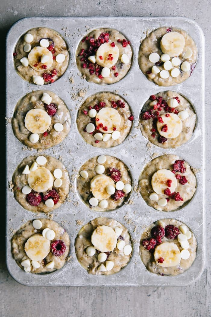 Muffin pan filled with coconut muffin batter and topped with one slice of banana, white chocolate chips, and crumbly frozen raspberries.