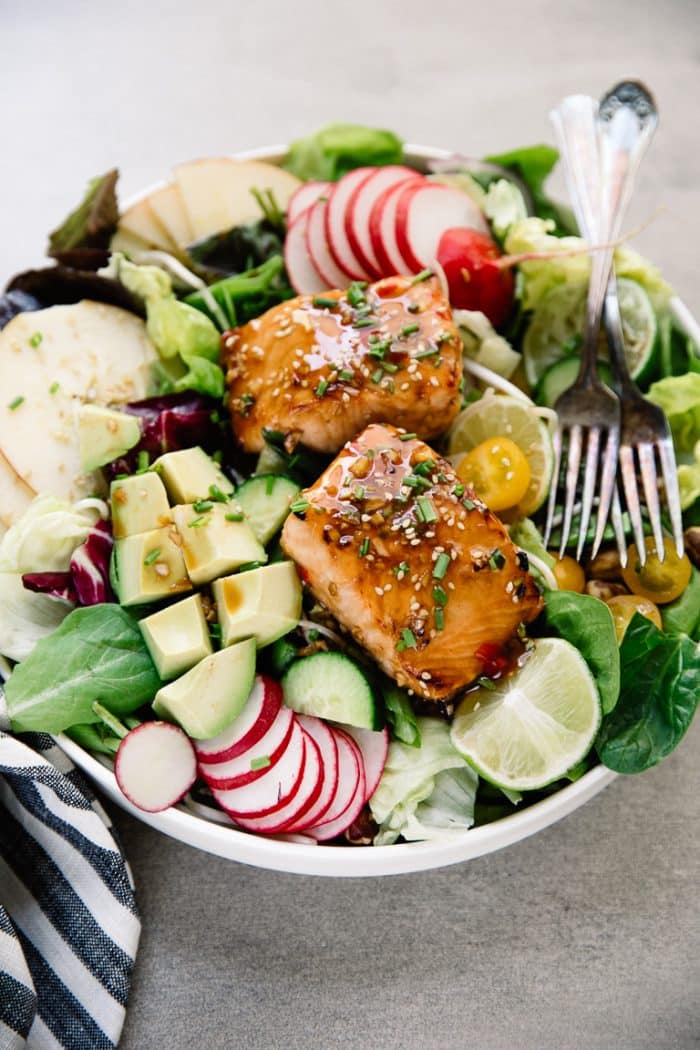 Large white salad bowl filled with apples, avocado, lettuce, tomatoes, radish, and topped with two cooked salmon fillets.