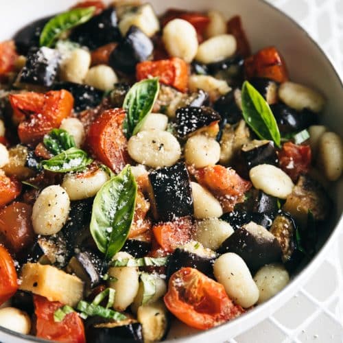 Close up image of a large white nonstick skillet filled with potato gnocchi, roasted tomatoes, eggplant, fresh basil, and parmesan cheese.