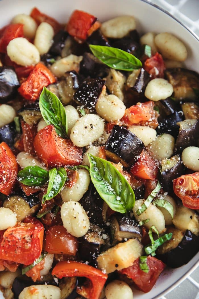 Close up image of a large white nonstick skillet filled with potato gnocchi, roasted tomatoes, eggplant, fresh basil, and parmesan cheese.