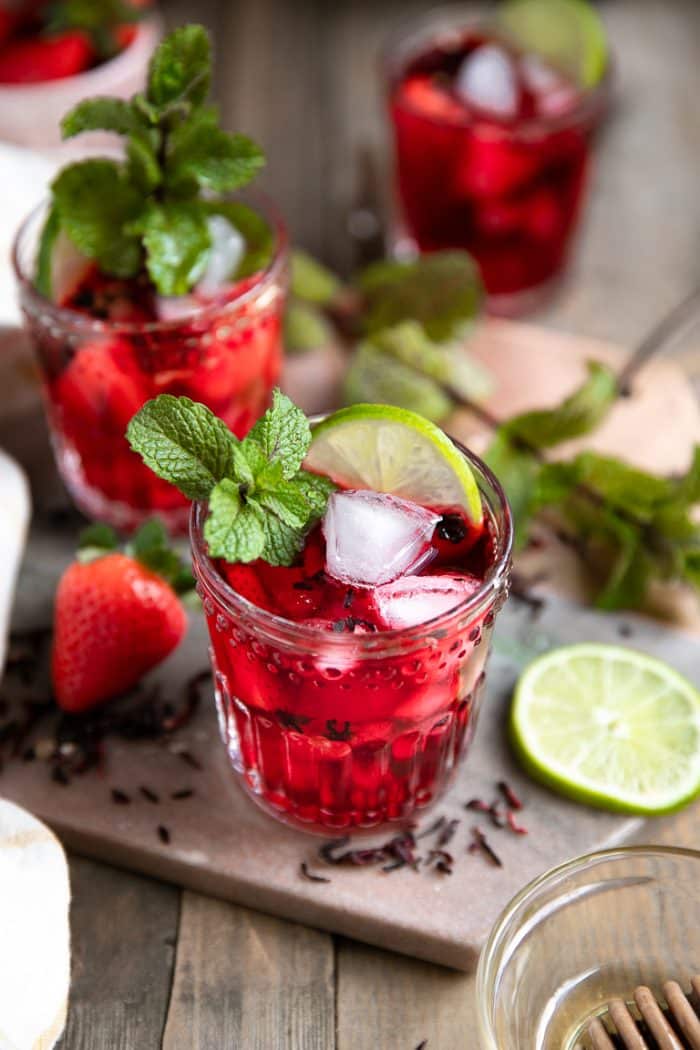three glasses of icy cold strawberry honey hibiscus tea topped with fresh mint, chopped strawberries, and sliced lime