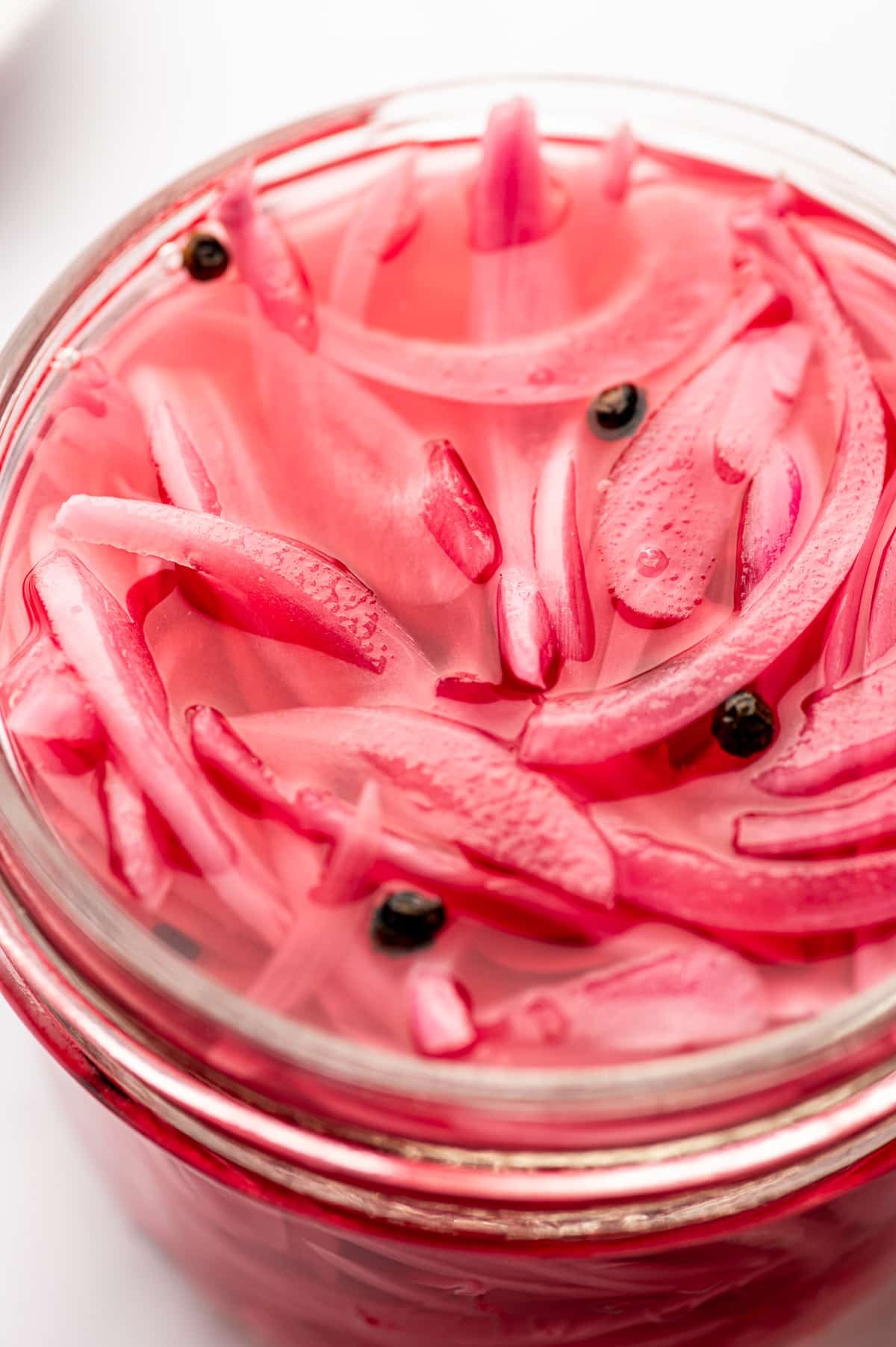 Extreme close-up of bright pink pickled red onion slices and black peppercorns in a clear glass jar, showing the glossy brine and thin onion layers.