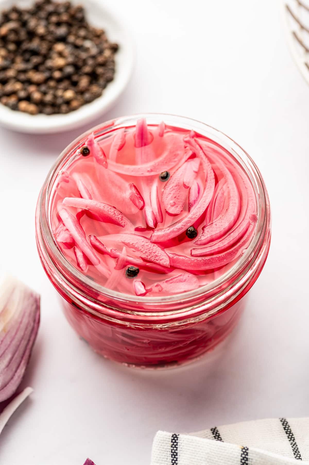 Overhead close-up of bright pink pickled red onions in a clear glass jar with black peppercorns, next to a small white bowl of peppercorns, a piece of red onion, and a white towel with black stripes.