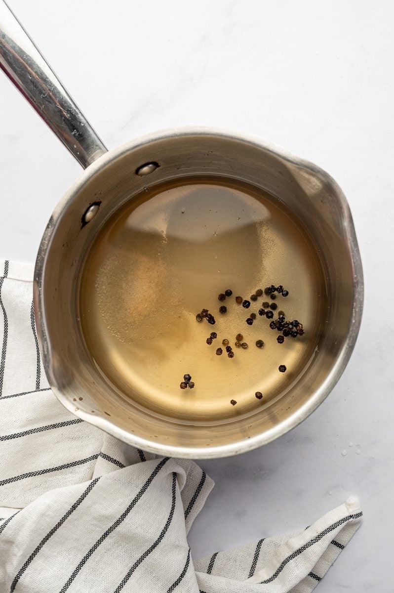 A small stainless steel saucepan filled with pickling brine, black peppercorns, sugar, and vinegar on a white marble surface with a white towel with black stripes.