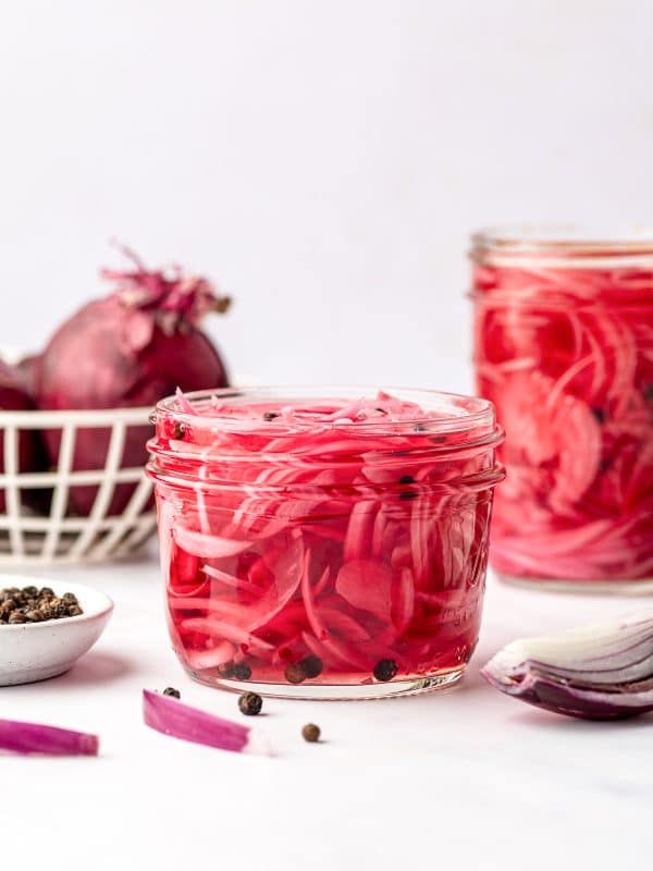 A clear glass jar filled with bright pink pickled red onions and black peppercorns, with a second jar in the background, whole red onions in a cream wire basket, a small white bowl of peppercorns, and sliced red onion on a white surface.