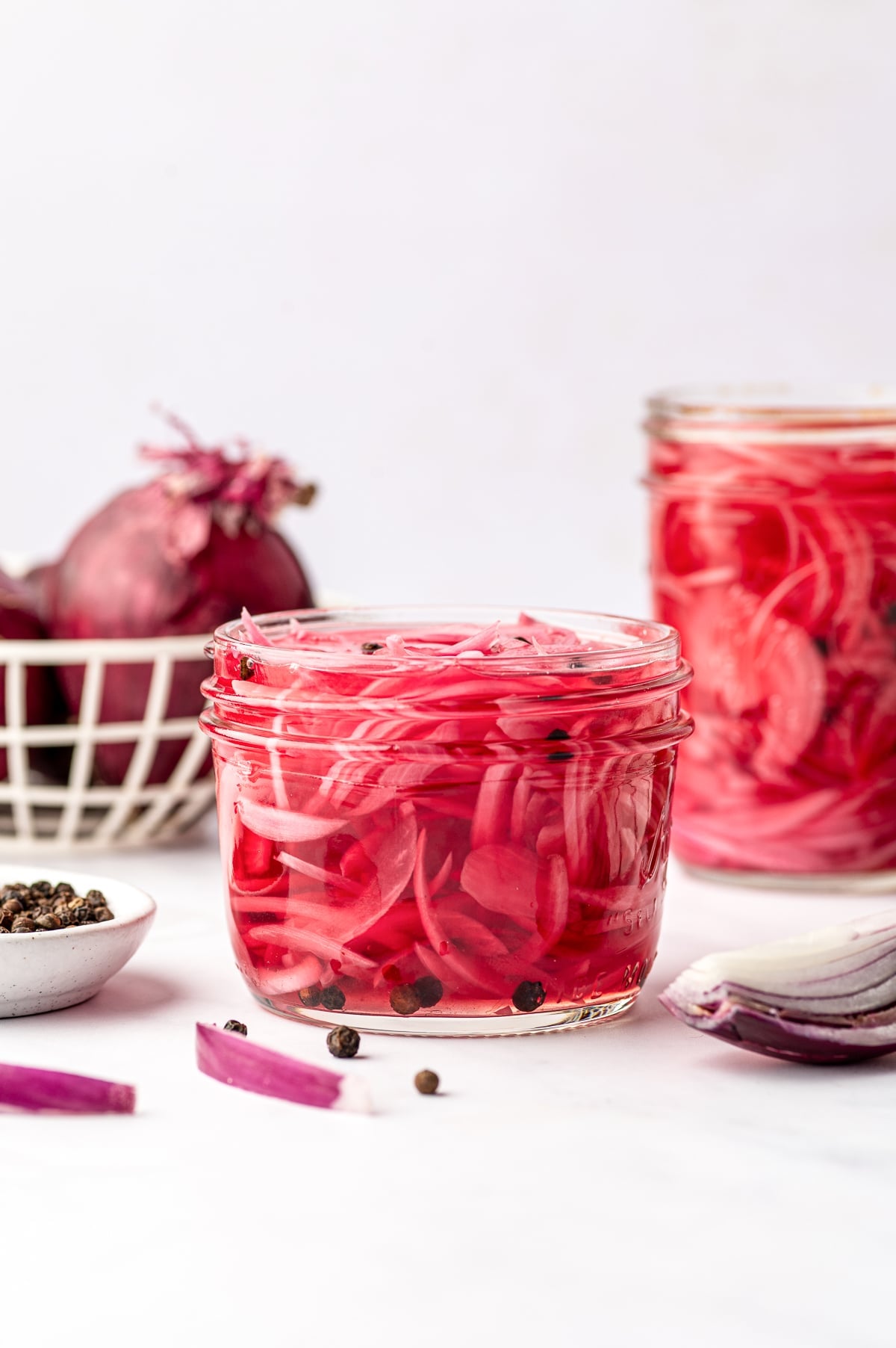 A clear glass jar filled with bright pink pickled red onions and black peppercorns, with a second jar in the background, whole red onions in a cream wire basket, a small white bowl of peppercorns, and sliced red onion on a white surface.
