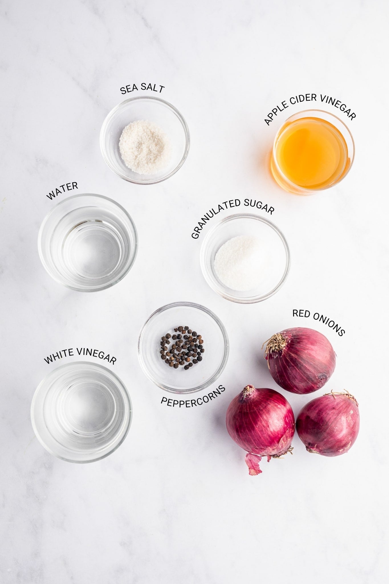 Overhead ingredient flat lay for pickled red onions with text labels for sea salt, apple cider vinegar, water, granulated sugar, red onions, white vinegar, and peppercorns arranged in small clear glass bowls on a white marble surface.