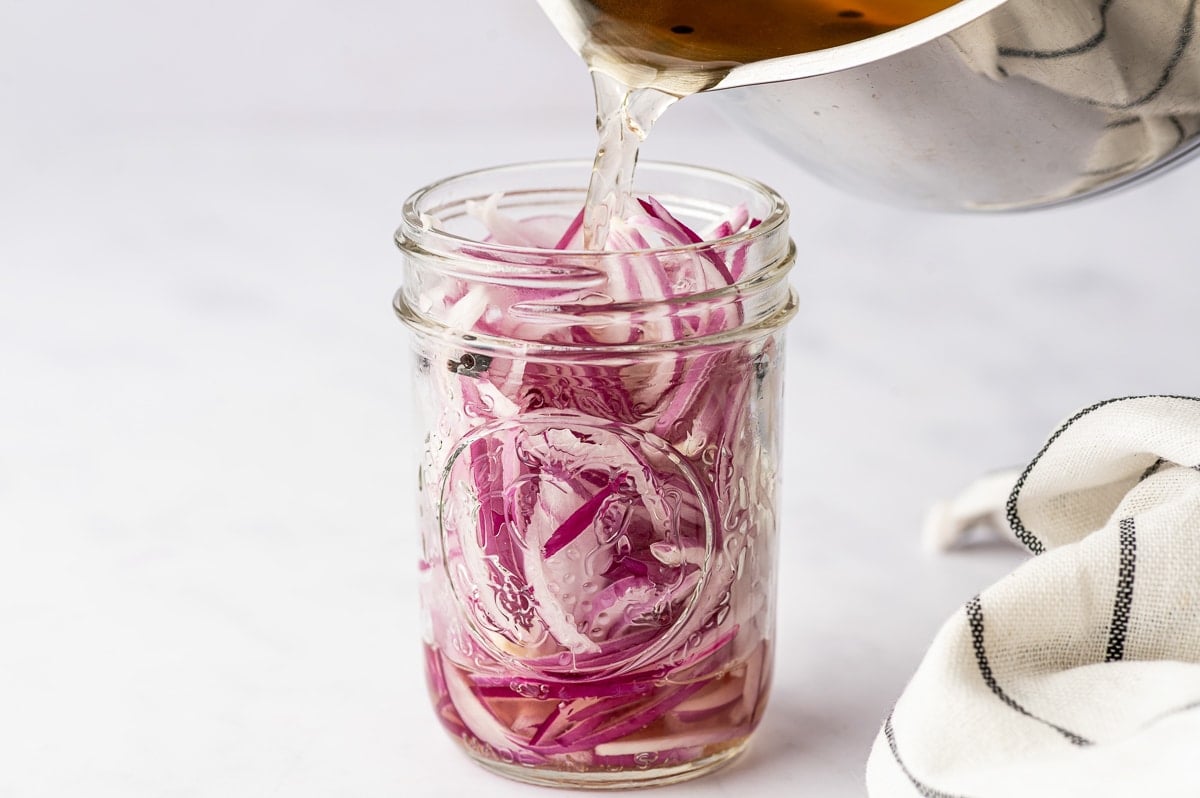 Warm pickling brine being poured from a stainless steel saucepan into a clear glass mason jar filled with thinly sliced red onions and black peppercorns, with a white towel with black stripes nearby.