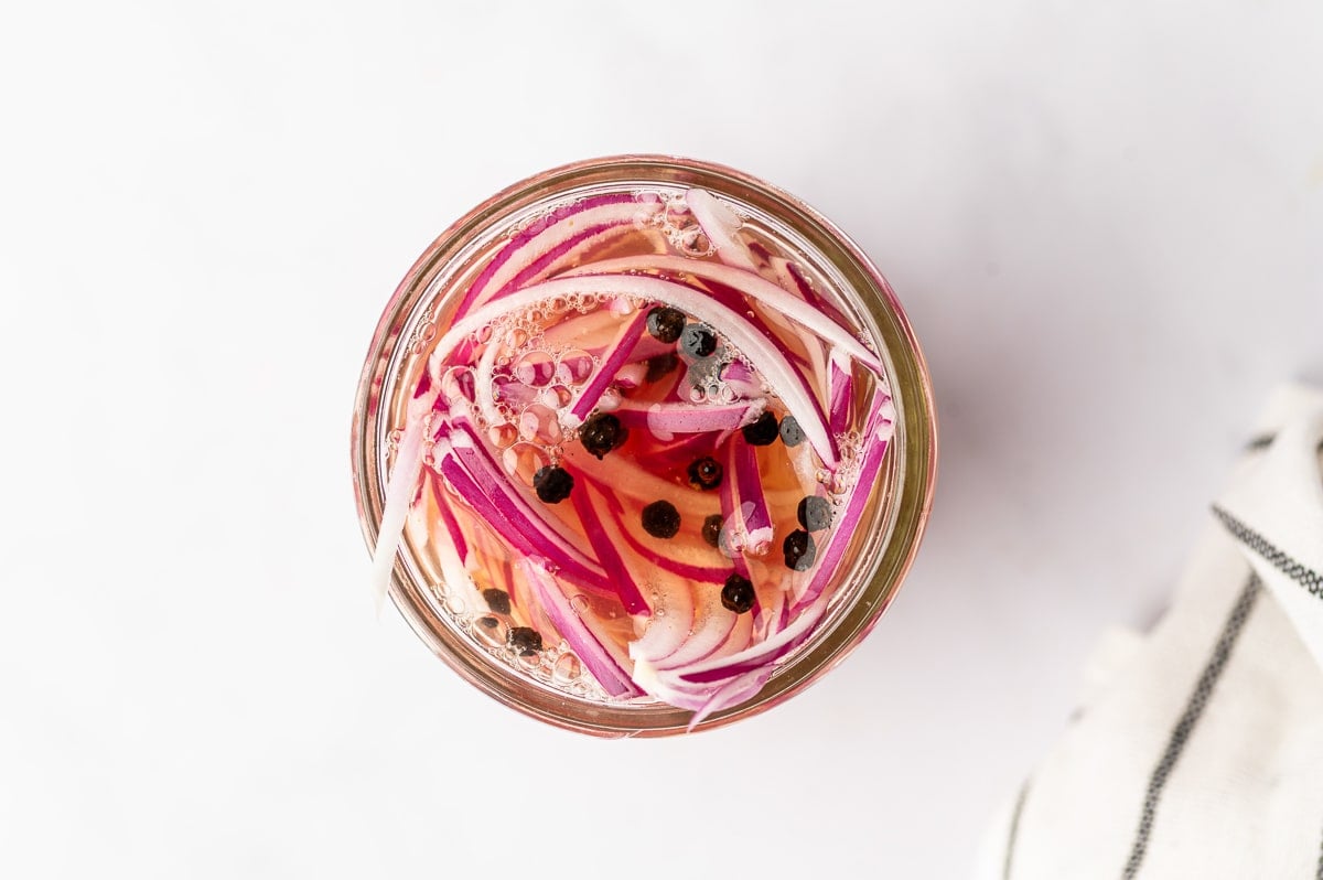 Overhead view of thinly sliced red onions and black peppercorns submerged in pale pink pickling brine inside a clear glass mason jar on a white surface.