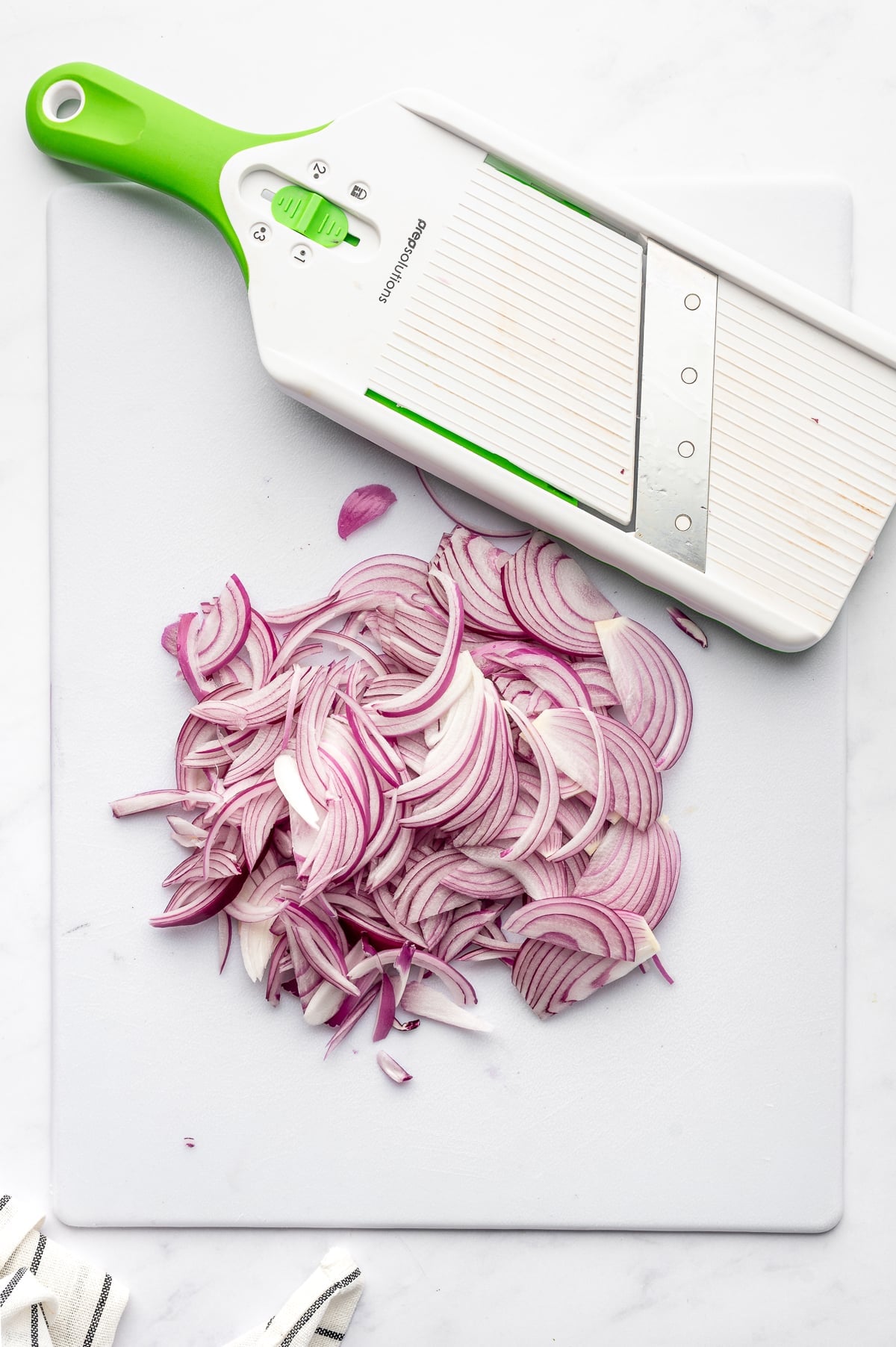 Thinly sliced red onions piled on a white cutting board beside a white and green mandoline slicer, showing evenly cut onion slices for quick pickled red onions.