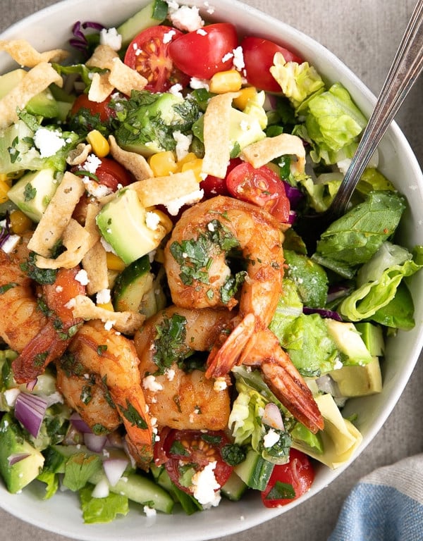 Large white bowl filled with chopped romaine, cherry tomatoes, cucumber, green onion, red onion, corn, avocado, and crumbled cheese, topped with plump tail-on cooked shrimp and drizzled with cilantro lime vinaigrette.