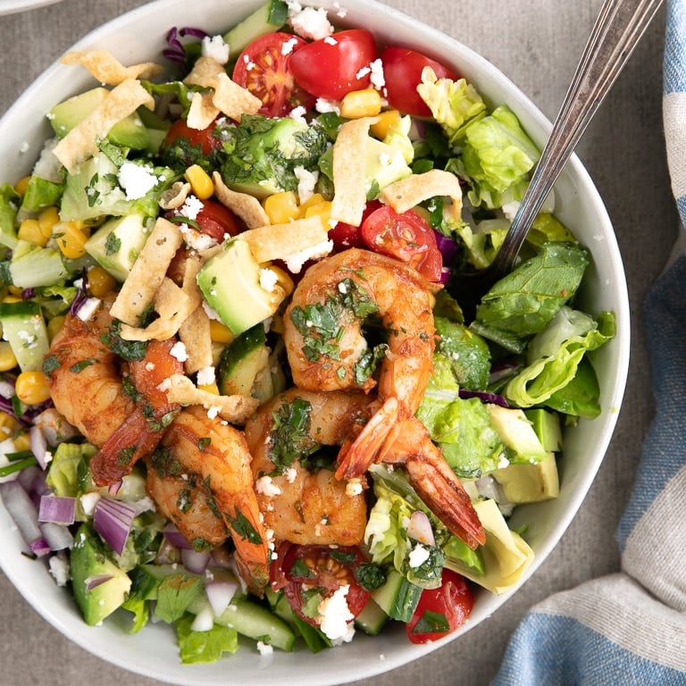 Large white bowl filled with chopped romaine, cherry tomatoes, cucumber, green onion, red onion, corn, avocado, and crumbled cheese, topped with plump tail-on cooked shrimp and drizzled with cilantro lime vinaigrette.