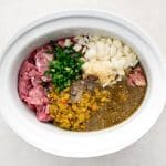 A large white slow cooker filled with chopped pork shoulder, minced onion, garlic, salsa verde, green chilies, jalapeño, and minced cilantro.