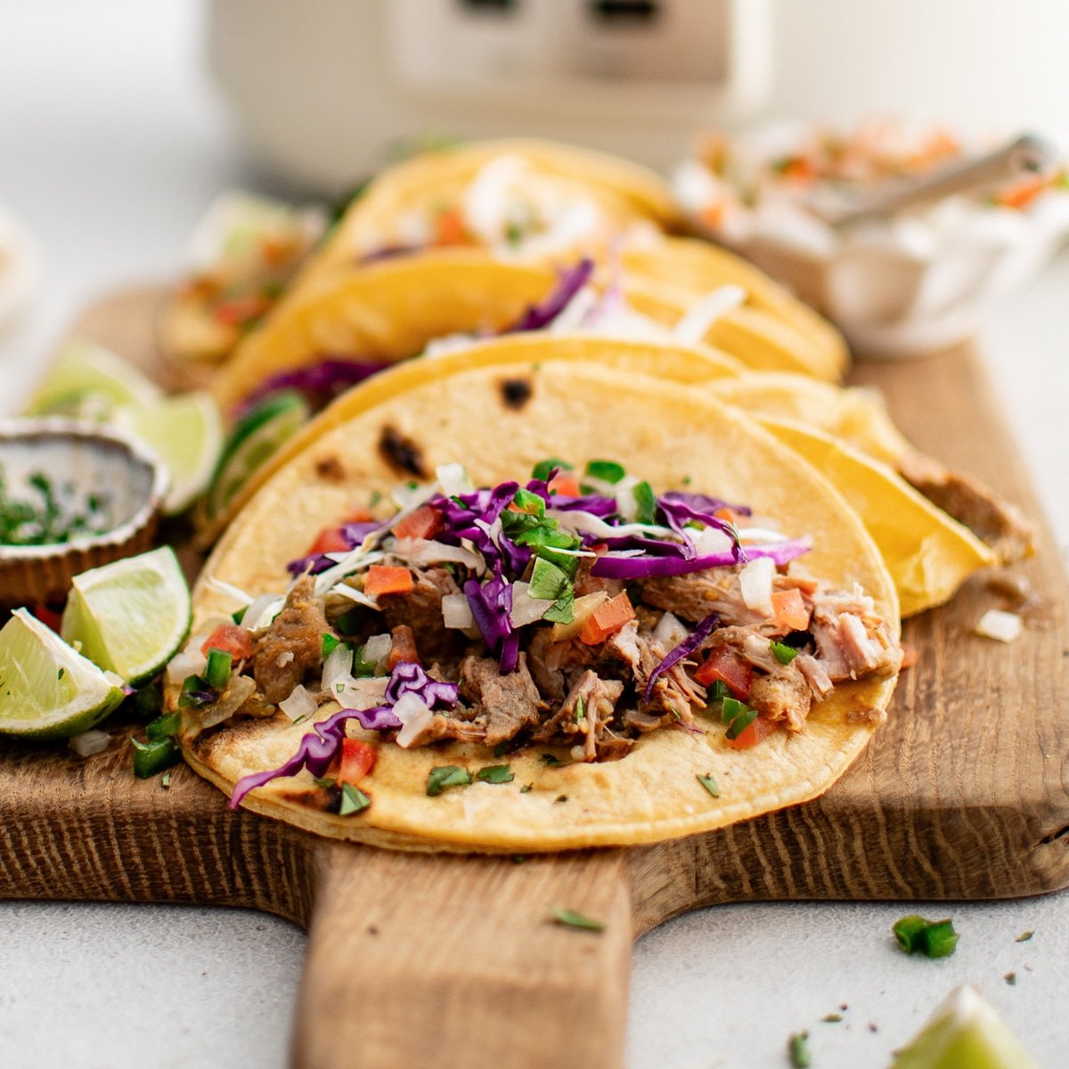 Corn tortillas filled with slow-cooked green chili pork, shredded red cabbage, and fresh pico de gallo presented on a wooden cutting board with a large white slow cooker in the background.