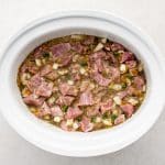 A large white slow cooker filled with mixed together chopped pork shoulder, minced onion, garlic, salsa verde, green chilies, jalapeño, and minced cilantro.