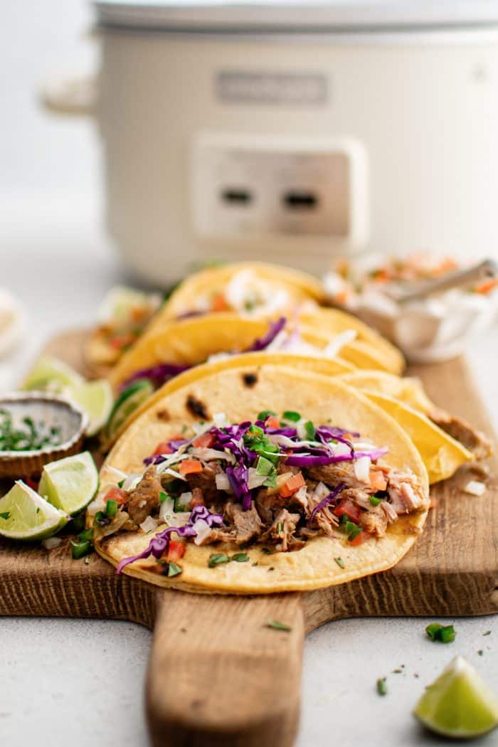 Corn tortillas filled with slow-cooked green chili pork, shredded red cabbage, and fresh pico de gallo presented on a wooden cutting board with a large white slow cooker in the background.