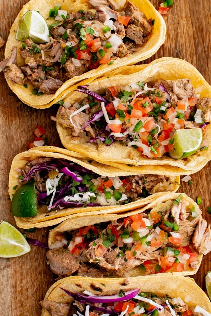 Corn tortillas filled with slow-cooked green chili pork, shredded red cabbage, and fresh pico de gallo with sliced limes on the side, all set on a large wooden cutting board.