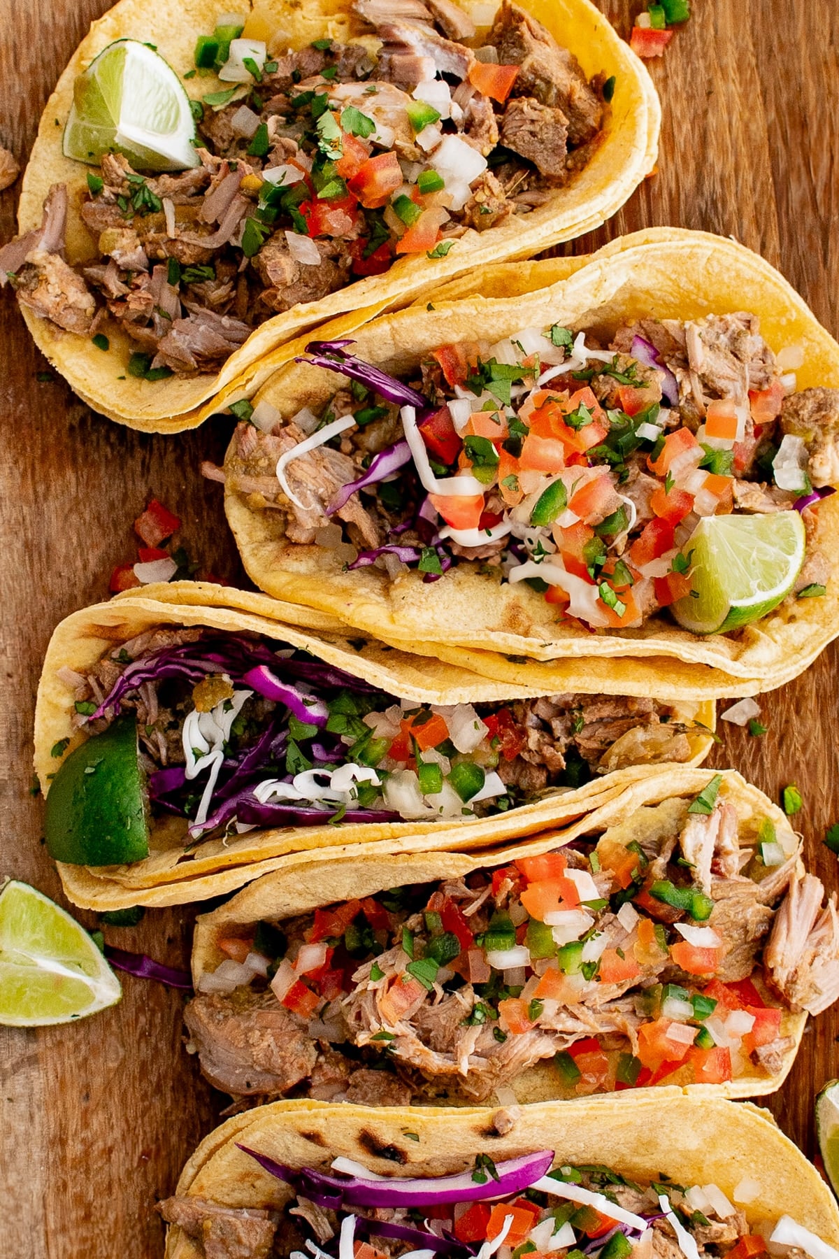 Corn tortillas filled with slow-cooked green chili pork, shredded red cabbage, and fresh pico de gallo with sliced limes on the side, all set on a large wooden cutting board.