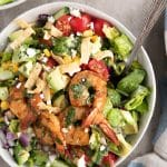 Large white bowl filled with chopped romaine, cherry tomatoes, cucumber, green onion, red onion, corn, avocado, and crumbled cheese, topped with plump tail-on cooked shrimp and drizzled with cilantro lime vinaigrette.