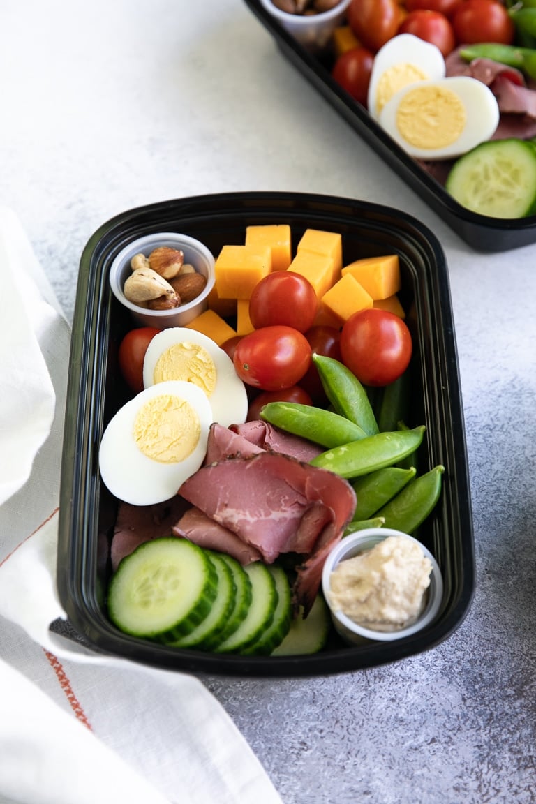 lunch meal prep tray with cheese, nuts, cherry tomatoes, sugar snap peas, cucumber, hummus, deli meat, and hardboiled egg