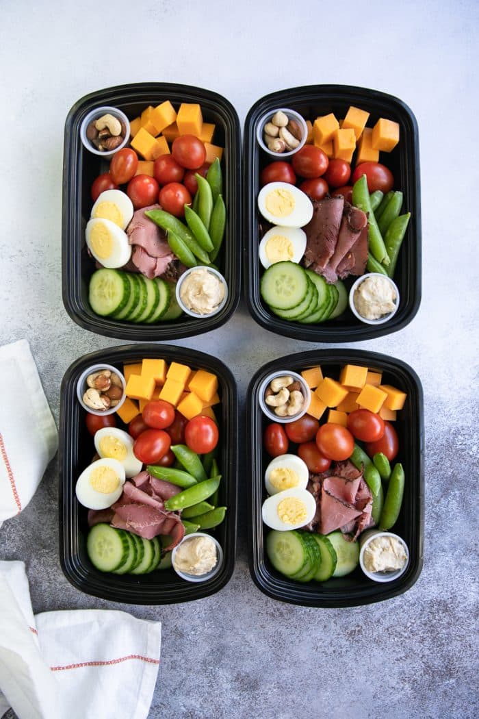 Four prepared high protein snack packs filled with healthy veggies and high protein snacks including cheese, cherry tomatoes, nuts, hummus, deli meat, and eggs.
