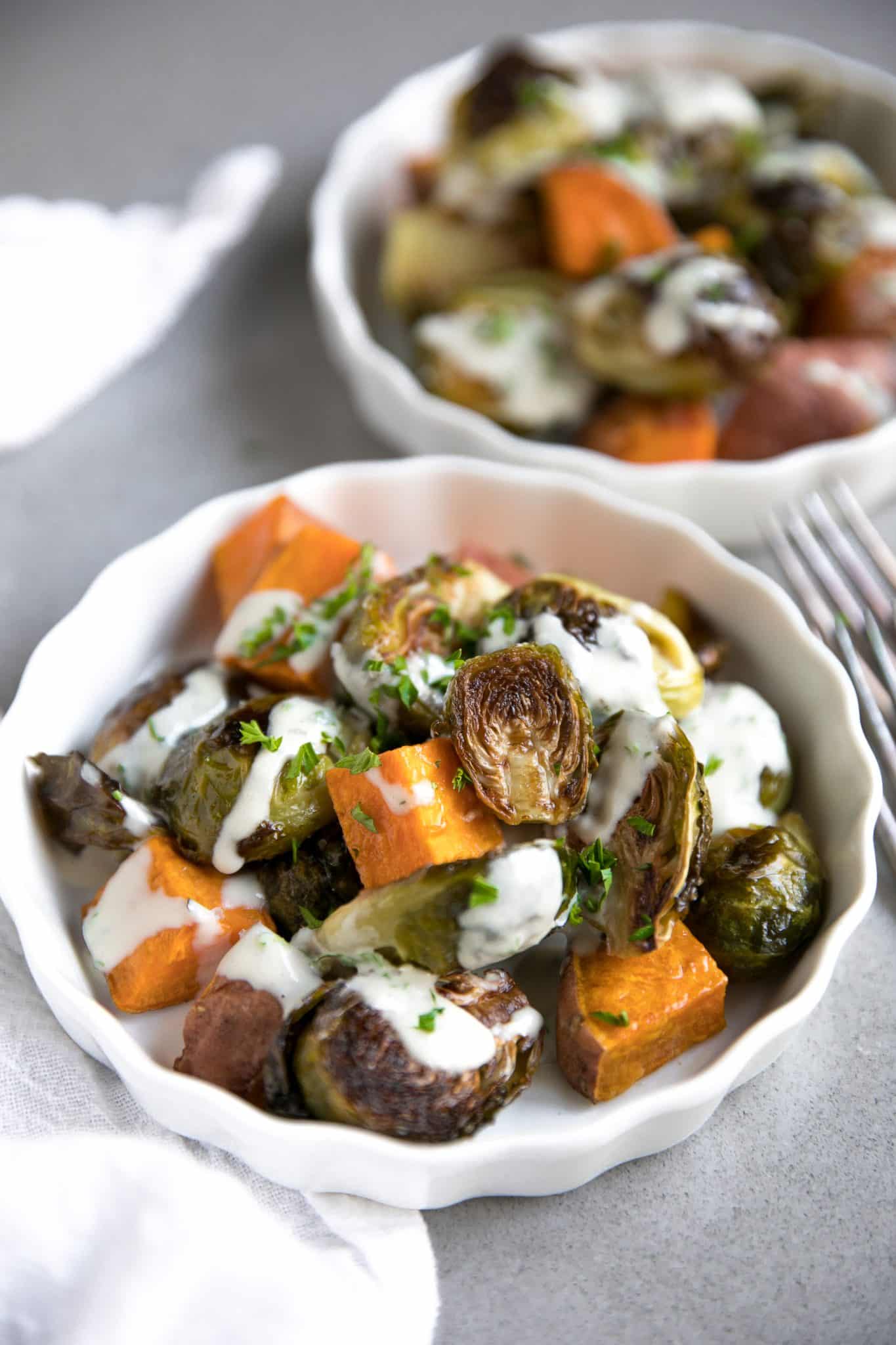 Small white plates filled with individual servings of oven-roasted Brussels sprouts and sweet potato cubes drizzled with tahini and chopped parsley.