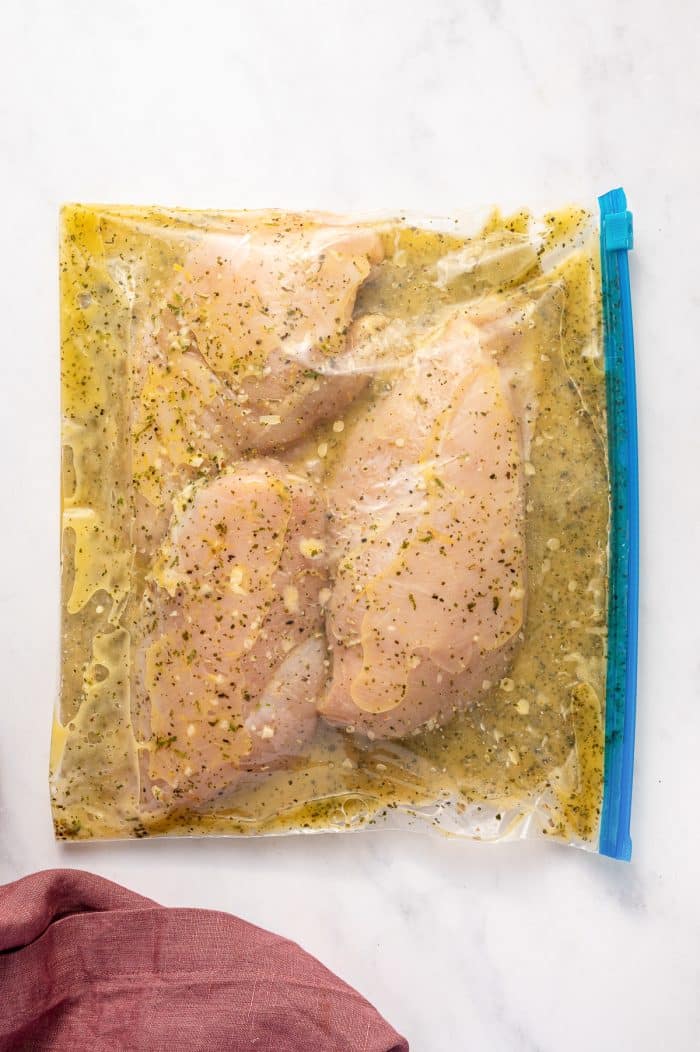 op-down flat lay of a sealed clear zip-top bag with a bright blue zipper strip filled with raw boneless skinless chicken breasts and citrus marinade on a white marble background. The pale yellow marinade is speckled with dried herbs and minced garlic and surrounds the pale pink chicken. A dusty rose pink linen napkin appears in the lower left corner.
