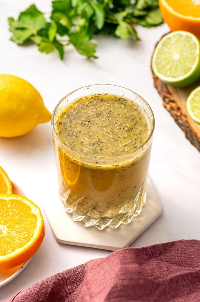 Angled close-up of a short textured glass filled with citrus chicken marinade on a pale hexagon coaster over a white marble background. The marinade is pale yellow with visible herbs and black pepper. Around the glass are a whole yellow lemon, halved limes, part of a halved orange, and fresh green parsley leaves. A dusty rose pink linen napkin is draped along the bottom right edge.