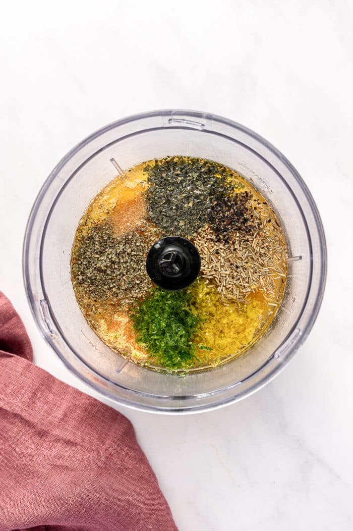 op-down view inside a clear food processor bowl filled with citrus chicken marinade ingredients on a white marble background. Visible ingredients include orange citrus juice, green lime zest, yellow lemon zest, dried rosemary, dried herbs, black pepper, salt, garlic powder, onion powder, and other seasonings arranged around the center blade. A dusty rose pink linen napkin is visible in the lower left corner.