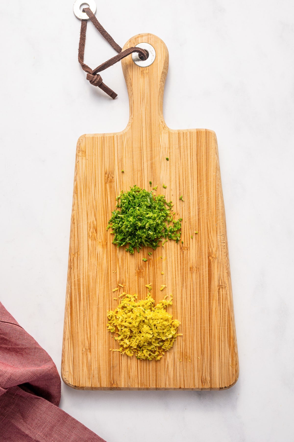 Top-down view of a light bamboo paddle cutting board on a white marble background with a brown leather hanging loop at the top, a small pile of finely grated green lime zest near the center, and a larger pile of finely grated yellow lemon zest near the bottom. A dusty rose pink linen napkin is draped into the bottom left corner.