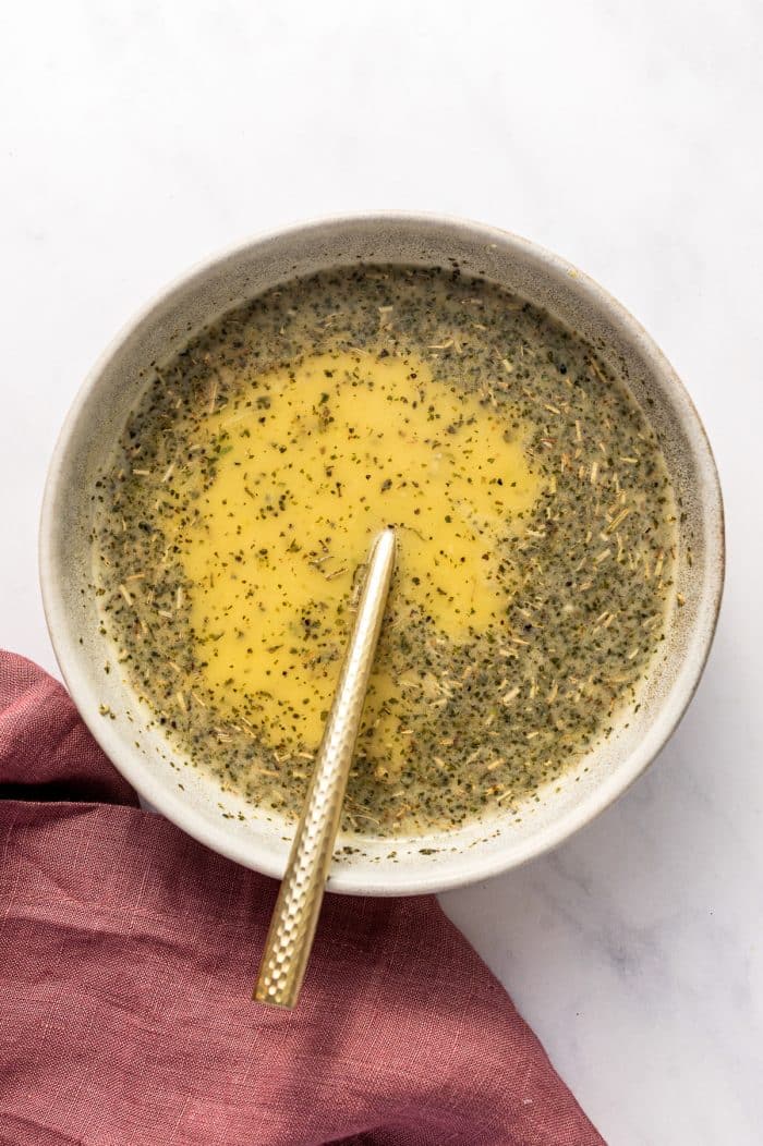 Top-down view of a speckled off-white ceramic bowl filled with pale yellow citrus chicken marinade flecked with dried herbs and seasonings on a white marble background. A gold spoon with a textured handle rests in the bowl, and a dusty rose pink linen napkin sits along the lower left edge. No text overlay.