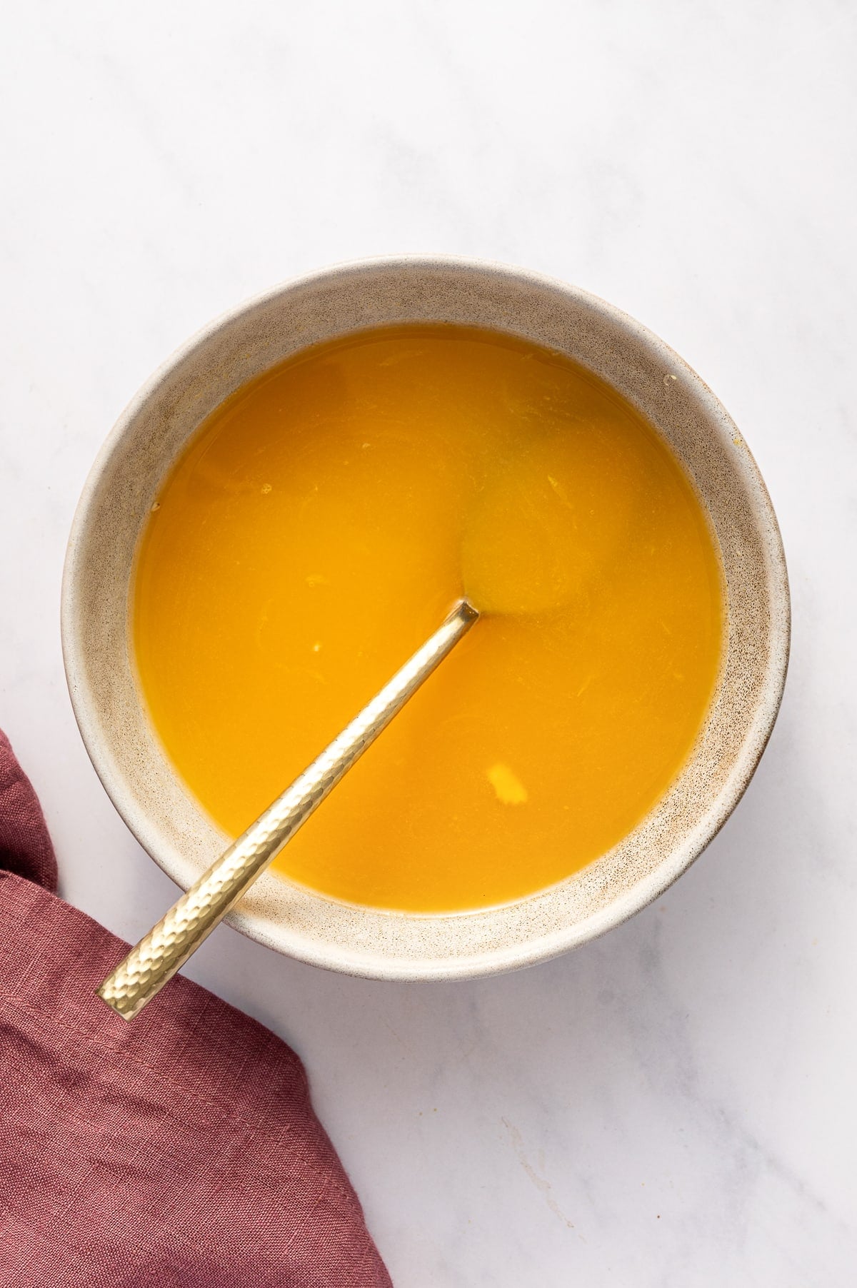 Top-down view of a speckled off-white ceramic bowl filled with bright orange citrus juice on a white marble background. A gold spoon with a textured handle rests inside the bowl. A dusty rose pink linen napkin sits in the lower left corner.