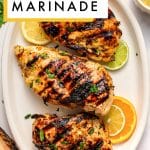 Vertical Pinterest pin for citrus chicken marinade showing three grilled chicken breasts on a large oval off-white platter with dark grill marks and chopped parsley garnish. Lemon, lime, and orange slices are tucked around the chicken, and a small off-white ramekin of pale yellow citrus marinade sits in the upper right corner. A white text box with a mustard yellow border overlays the top left corner and reads โCITRUS CHICKEN MARINADEโ in bold black letters. A rustic wood slice, fresh parsley, citrus slices, and a dusty rose pink linen napkin surround the platter on a white background. The text โthe forked spoonโ appears at the bottom.