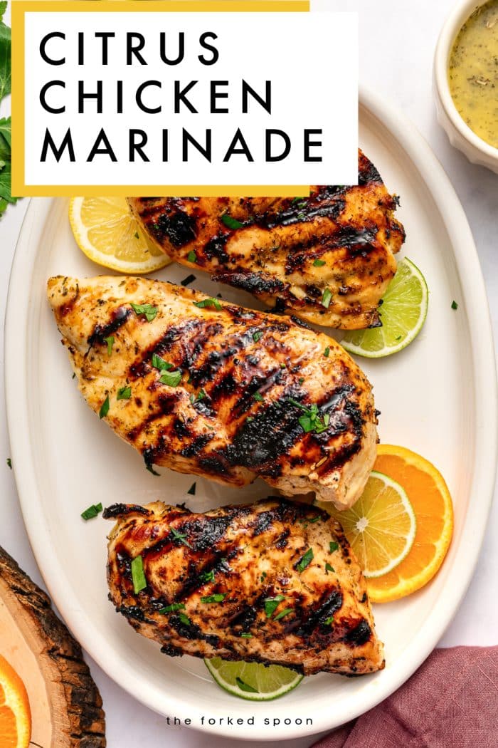 Vertical Pinterest pin for citrus chicken marinade showing three grilled chicken breasts on a large oval off-white platter with dark grill marks and chopped parsley garnish. Lemon, lime, and orange slices are tucked around the chicken, and a small off-white ramekin of pale yellow citrus marinade sits in the upper right corner. A white text box with a mustard yellow border overlays the top left corner and reads “CITRUS CHICKEN MARINADE” in bold black letters. A rustic wood slice, fresh parsley, citrus slices, and a dusty rose pink linen napkin surround the platter on a white background. The text “the forked spoon” appears at the bottom.